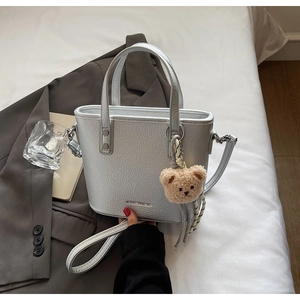 Silver grain leather bucket bag with top handles and plush bear charm on white linen