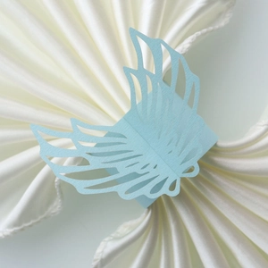 Detailed view of light blue laser-cut angel wing paper napkin rings on cream fabric