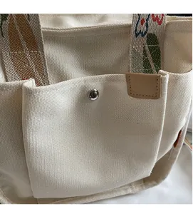 Detail of off-white canvas tote bag featuring silver metal snap button and embroidered strap