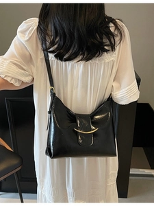 Minimalist black glossy shoulder bag with gold metal clasp worn by woman in white dress