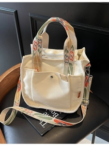 White canvas tote bag with floral embroidery straps and front pockets on a black chair
