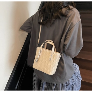 Dettaglio di borsa a tracolla beige intrecciata con doppi manici e ciondolo a cuore in metallo