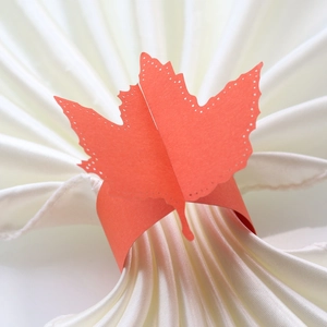 Detail of laser-cut paper maple leaf napkin ring in vibrant orange on white satin cloth