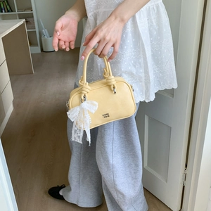Detail einer hellgelben Handtasche mit Knotengriff und weißem Spitzen-Schleifenanhänger
