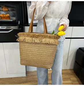 Detail of woven straw tote bag featuring brown leather handles and decorative fringe