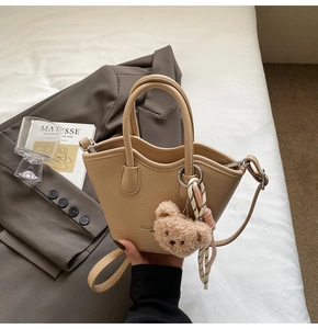 Détail du sac seau kaki avec breloque ours en peluche et ruban sur fond de literie neutre