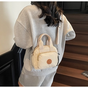 Detail der cremeweißen Nylon-Umhängetasche mit Logo und Doppelreißverschlüssen auf Holzstufen