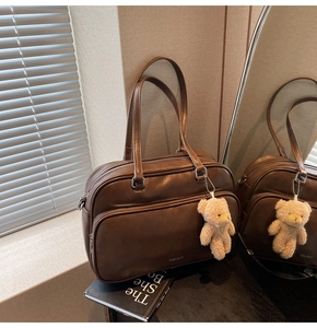 Detail of a large brown Boston bag with silver zippers and a plush bear charm on a desk