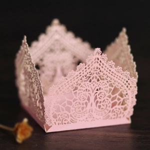 Intricate pink laser-cut lace cupcake wrapper with crown motif on dark wood background