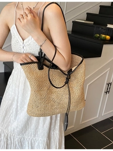 Large woven straw tote bag with black leather handles shown on shoulder in modern interior