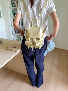 Pale yellow leather backpack with drawstring and metal buckles worn by a model in a room