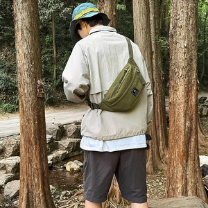 Detailed view of an olive green nylon sling bag worn by a model in an outdoor forest setting