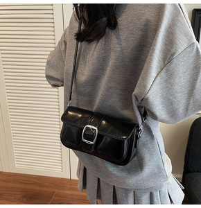 Detail of a black glossy crossbody bag with silver buckle worn by a model in casual grey