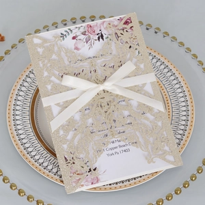 Detail of laser-cut glitter wedding invitation with floral insert and cream silk ribbon