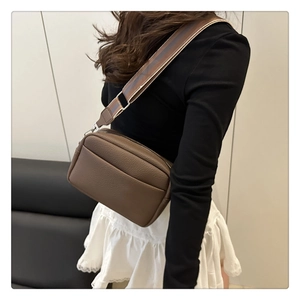 Close-up of brown litchi leather camera bag with wide striped fabric strap on a model