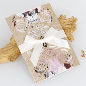 Detail of laser-cut glitter gold wedding invitation with floral insert and white silk bow