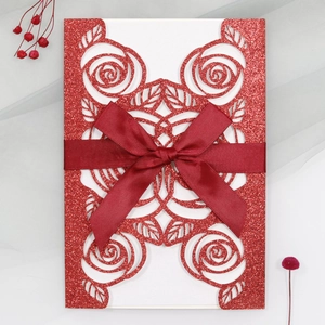 Close-up of laser-cut glitter paper wedding invitation with rose hollow and red ribbon bow