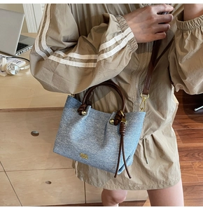 Dettaglio di una borsa a secchiello in denim azzurro con manici in pelle marrone e logo