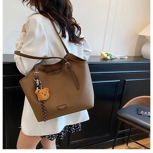 Brown litchi grain leather tote bag with plush bear charm worn by model in white suit
