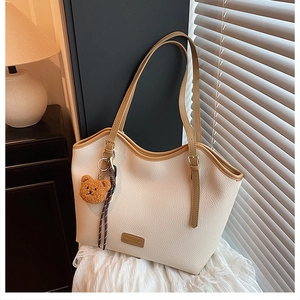 Close-up of a cream tote bag with brown straps, metallic logo, and cute bear charm