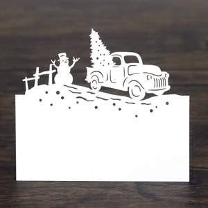 Detail of white laser-cut Christmas place card with snowman and truck on wooden table