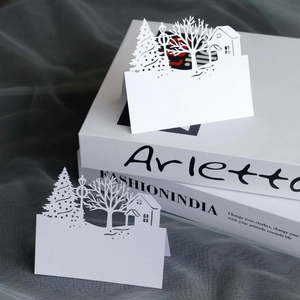 Close-up of laser-cut Christmas table card showing a tree and house in white pearlescent paper