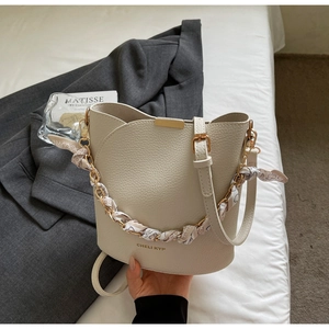 Sac seau chic en cuir crème avec poignée chaîne et foulard en soie sur un lit blanc