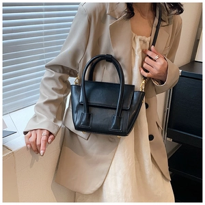 Detail of a black trapezoid crossbody bag with gold hardware worn with a beige blazer