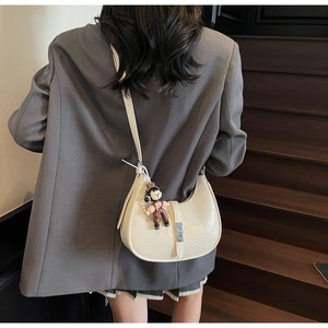 Chic off-white crescent crossbody bag featuring glossy crinkled leather and cute doll pendant