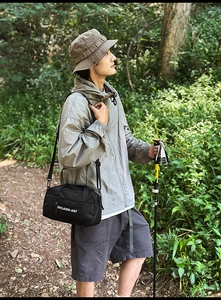 Outdoor scene showing a black nylon crossbody bag with logo and hiking stick in forest