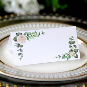 Detail view of a white folded wedding place card with elegant white rose and green leaf decor