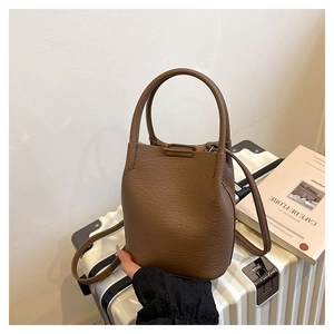 Brown litchi grain leather bucket bag shown on a white suitcase with travel magazines