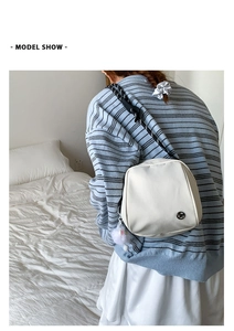 White crossbody bag with striped wide strap and fluffy keychain detail on a soft bed
