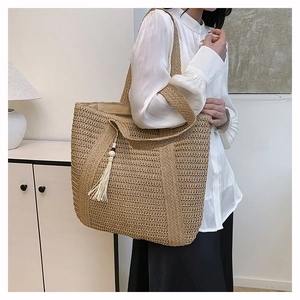Detail of beige woven straw tote bag with tassel pendant on model's shoulder