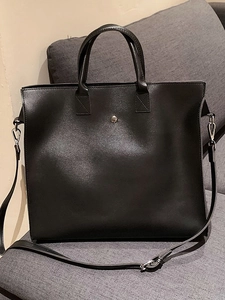 Minimalist black leather tote bag with silver buckle and adjustable shoulder strap on sofa