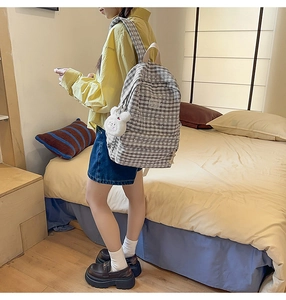 Gray plaid backpack with cute bunny charm shown on person in a cozy bedroom setting