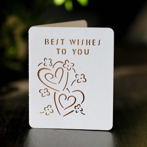 Detail of white laser-cut card with Best Wishes text and heart motifs on wood