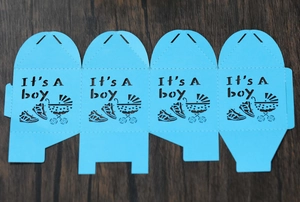 Flat lay of laser-cut light blue candy box with 'It's A boy' text and baby carriage pattern