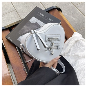 Detail of heart-shaped crossbody bag with dual zippers and silver chain on a dark bench