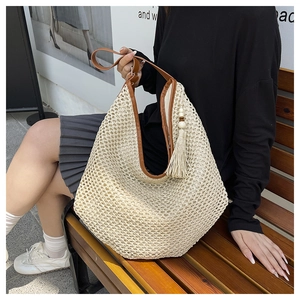 Detail of beige straw hobo bag with brown leather trim and tassel on a wooden bench