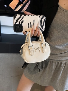 Mini cream bowling handbag with metal bow detail and dual handles shown for size scale
