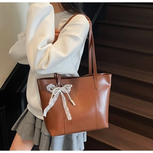 Detail of a brown matte leather tote bag featuring a white lace bow and pearl pendant