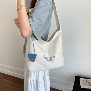 Close-up of cream canvas tote bag with blue C'est lavie embroidery and leather charm