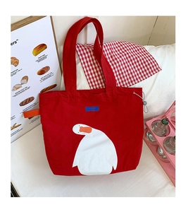 Detail showing red canvas tote bag with white duck illustration on a cozy bed background