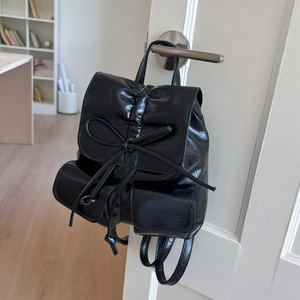 Black synthetic leather backpack with pleated design and bow detail hanging on a white door