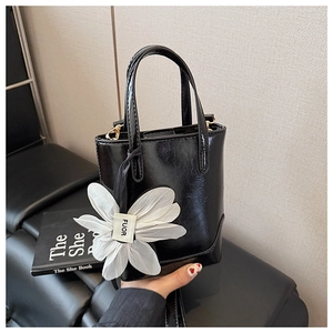 Detail of glossy black bucket bag with a white flower charm on a leather sofa