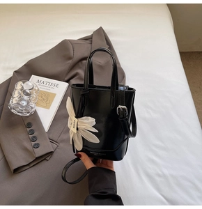 Détail d'un sac seau en cuir verni noir avec un pendentif fleur blanche et une bandoulière