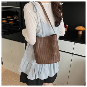 Detail of a dark brown woven leather bucket bag worn by a model in a cozy kitchen setting