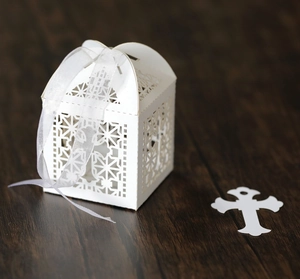 Detailed laser-cut white cross favor box with sheer ribbon on dark wood background