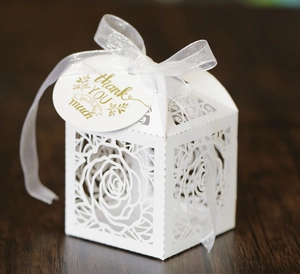 Laser-cut white wedding favor box with rose patterns and thank you tag on wood surface